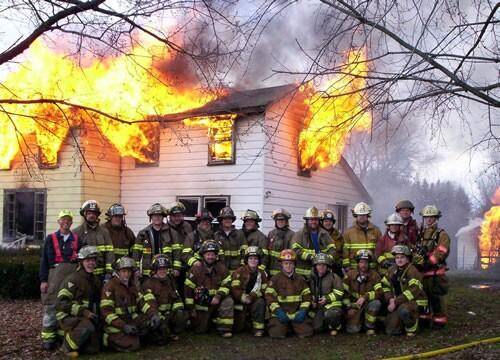 a group of firemen posing for a photo in front of a burning building